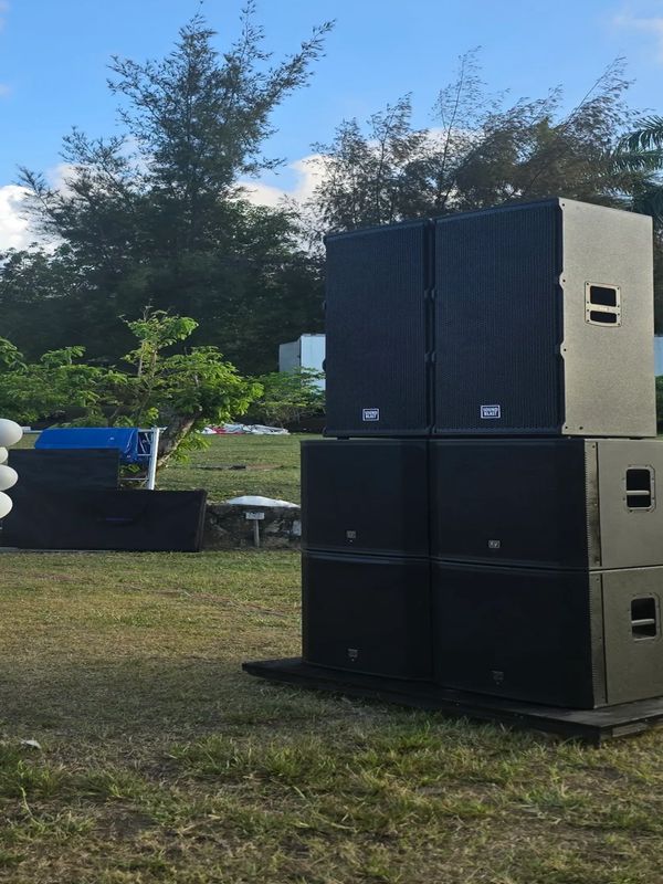 Stacked black speakers set outdoors on grass under a blue sky.