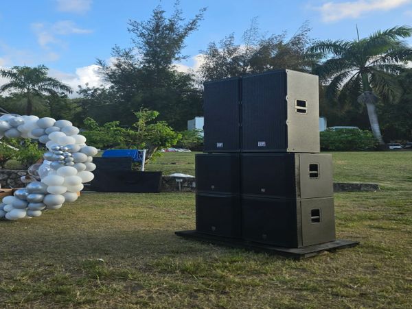 Stacked black speakers set outdoors on grass under a blue sky.
