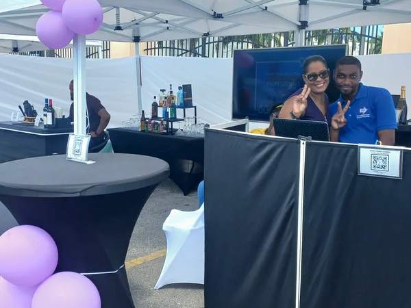Two people posing with peace signs at an outdoor booth with drinks and purple balloons.