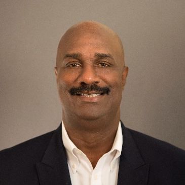 Smiling man in a dark blazer and white shirt against a plain background.