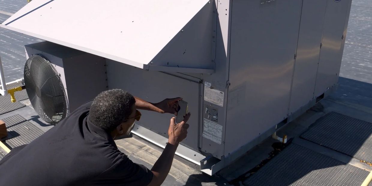 Technician inspecting and photographing rooftop HVAC unit on a sunny day.