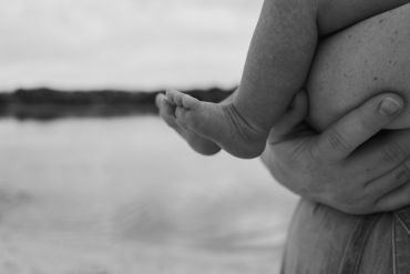 A person gently holding a baby's leg near a body of water in black and white.