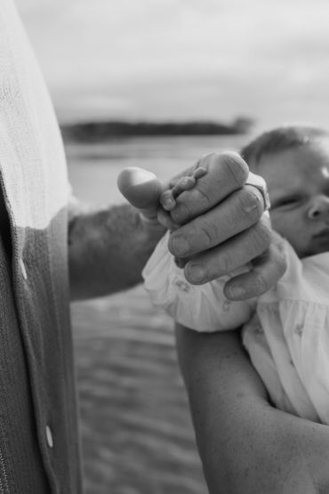 Black and white photo of an adult holding a baby's hand gently near water.