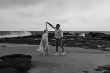 Couple dancing joyfully on a rocky beach under cloudy skies.