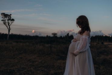 Pregnant woman in a flowing white dress stands at dusk in a field.