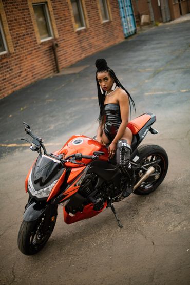 Model on motorcycle wearing a black stretch vinyl bodysuit with lighter earrings.