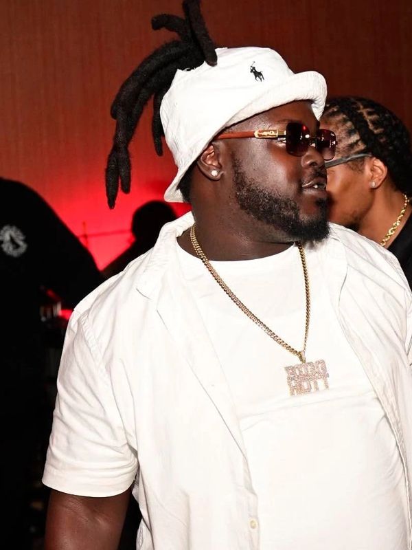 Man wearing white outfit, sunglasses, and a hat with dreadlocks, accessorized with a gold chain.