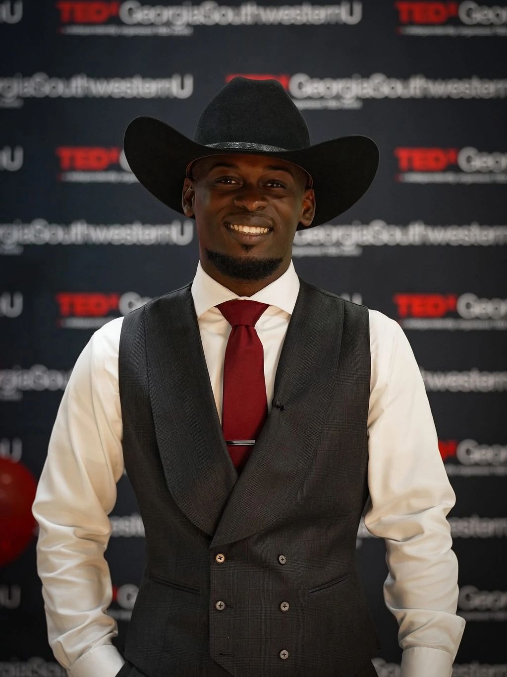 A man in a black cowboy hat, gray vest, white shirt, and red tie smiling at a TEDx event.