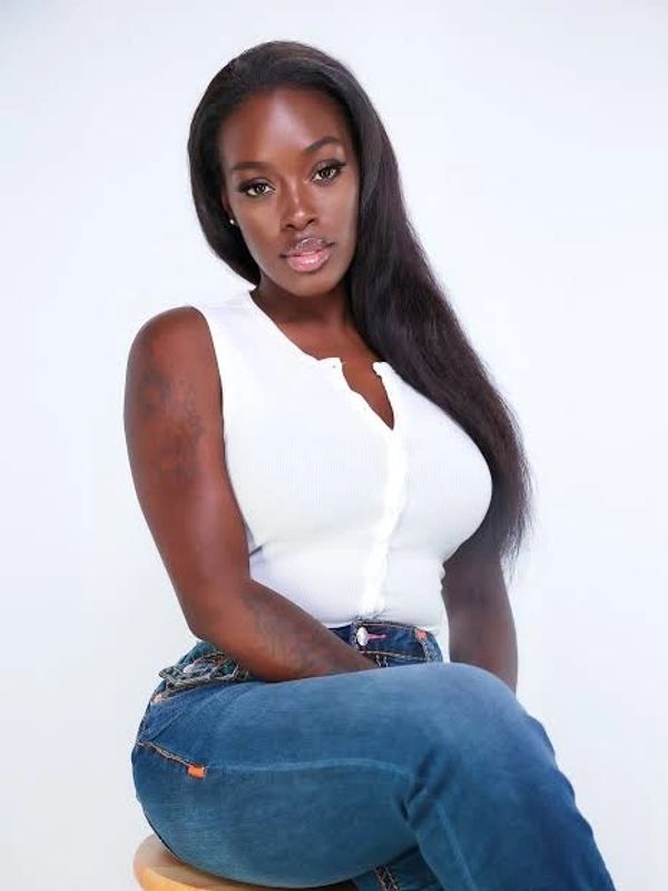Confident woman in white top and blue jeans sitting on a stool against a plain backdrop.