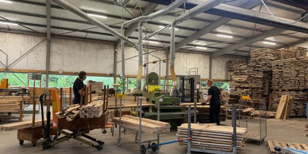 Twee mannen werken in een houtzagerij met stapels hout en machines.
