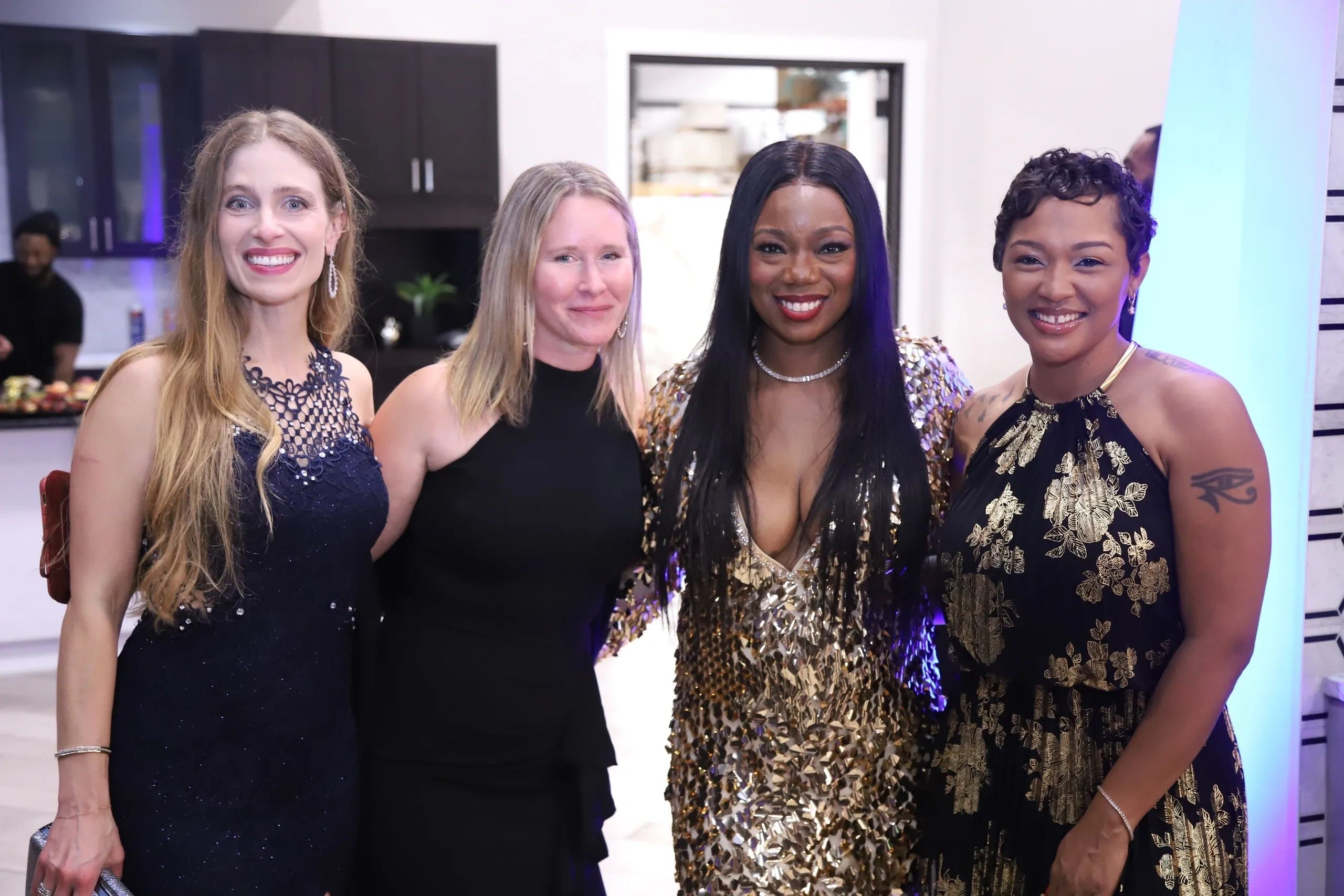 Four women dressed elegantly, smiling together at an indoor event.