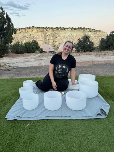 A woman smiling outdoors with white crystal singing bowls arranged on a blanket.