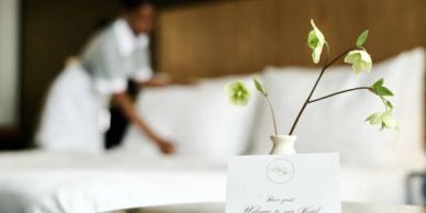 A hotel room with a welcome card and a maid making the bed in the background.