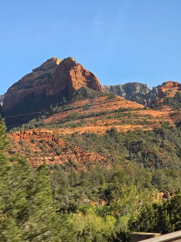 Red rock mountain under clear blue sky with surrounding green trees.