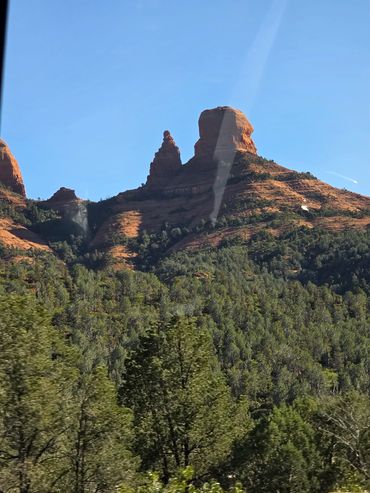 Red rock formations rise above dense green forest under clear blue sky.