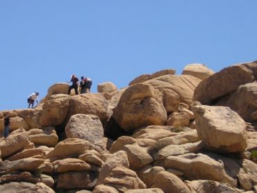 People climbing large rocky boulders under a clear blue sky.