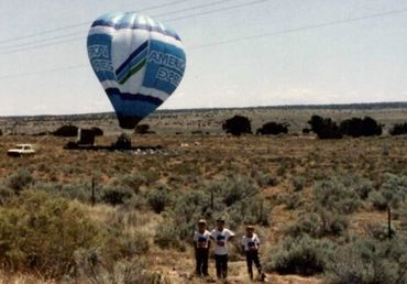 Three people stand in a field near a hot air balloon preparing to take off.