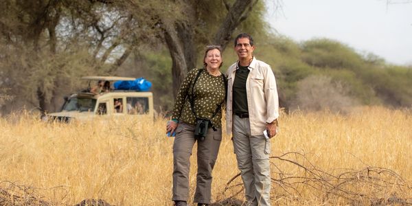 A smiling couple stands in dry grassland with a safari vehicle in the background.