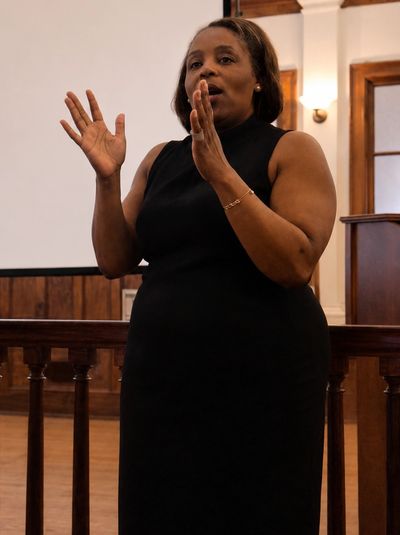 Woman in black dress speaking expressively indoors.
