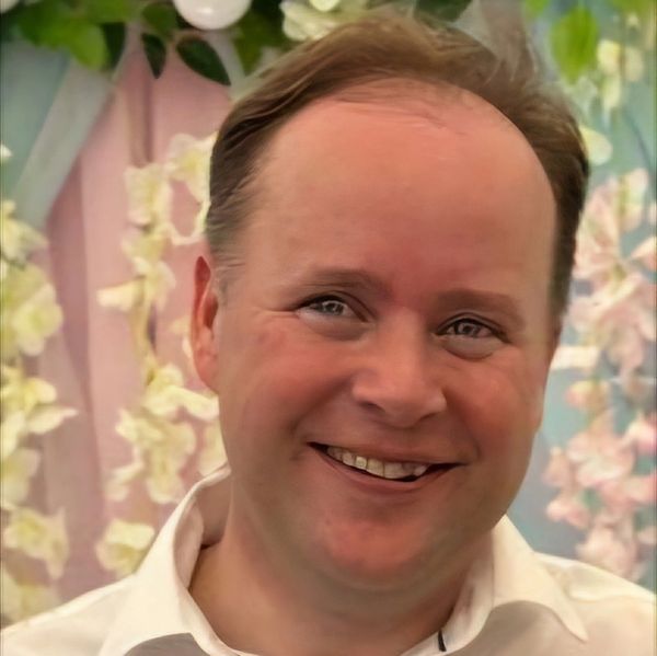 Smiling man in a white shirt with floral background and hanging lights.