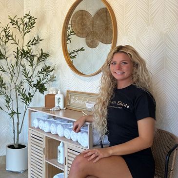 Woman with long blonde hair sitting beside a shelf with rolled towels and decor.