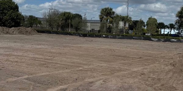 Cleared and leveled construction site with some trees and buildings in the background.