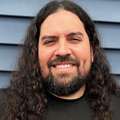 Smiling man with long curly hair and beard in front of blue siding.