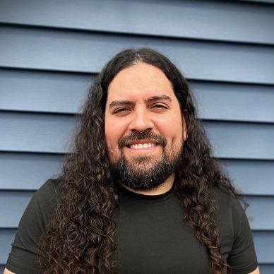 Smiling man with long curly hair and beard in front of blue siding.