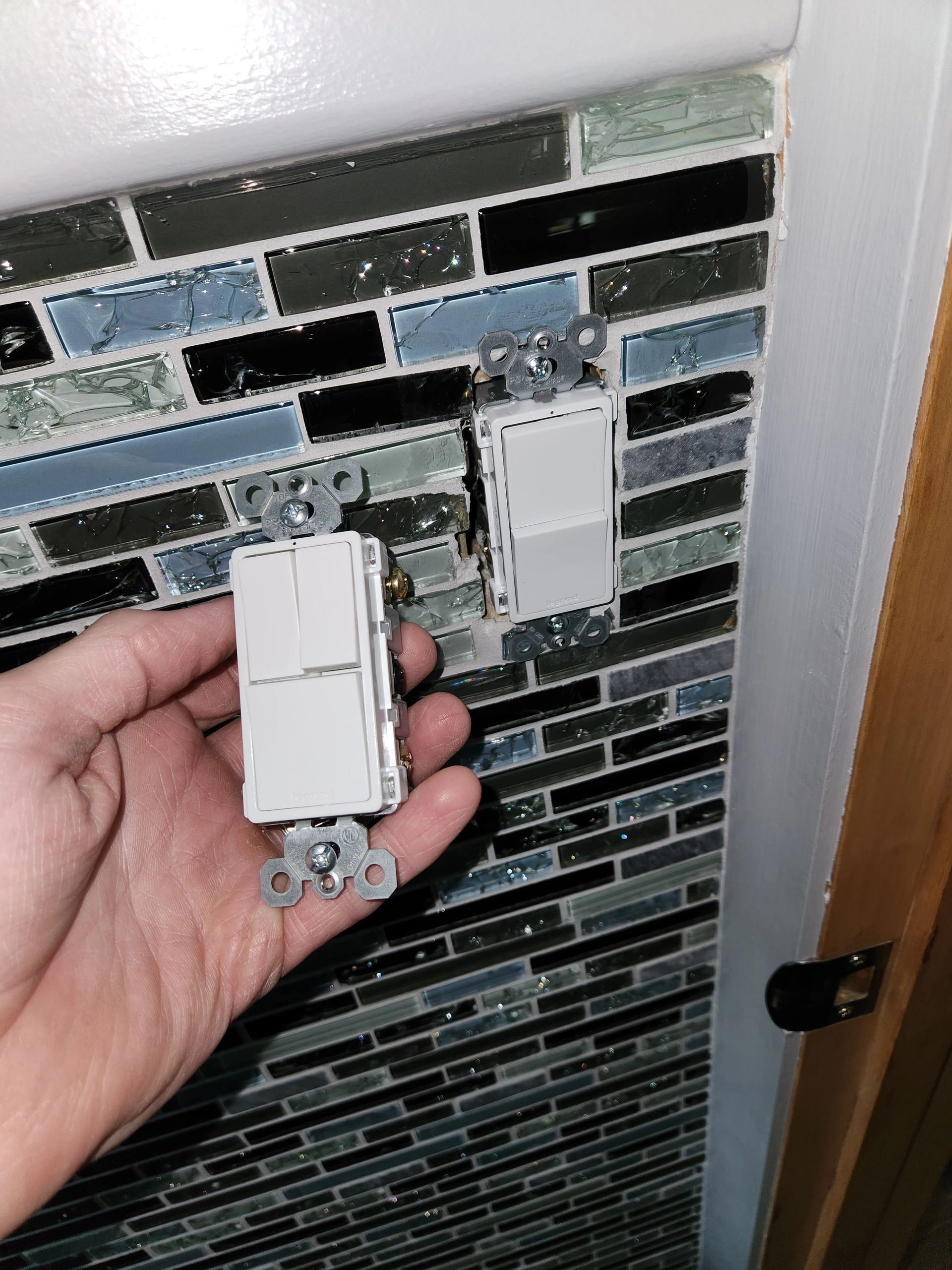 Hand holding a light switch near an installed switch on a tiled wall.