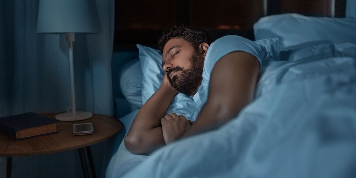 Man sleeping in bed, deeply dreaming, with book on side table to understand why do we dream.