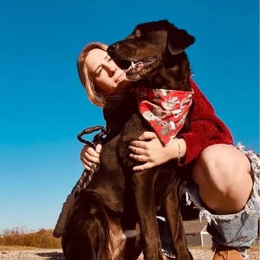 A woman embraces her black dog wearing a red bandana under a clear blue sky.