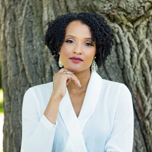 Confident woman with curly hair in white outfit poses thoughtfully by a tree.