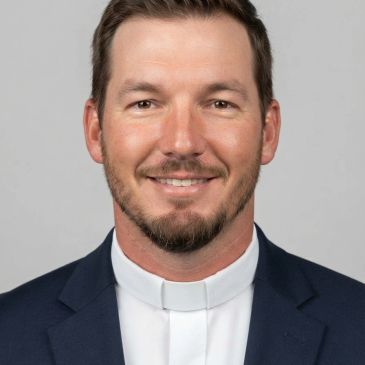 Smiling man wearing clerical collar and dark blazer.