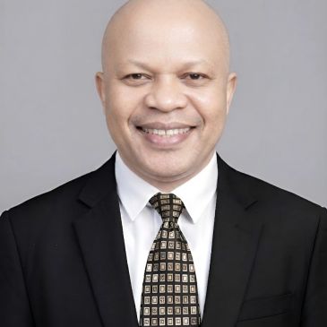 Man in black suit smiles against grey background.