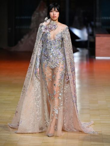 Model walks runway in sheer floral-embellished gown with cape.