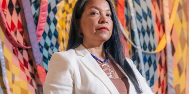 A confident woman in a white blazer stands before colorful patterned banners.