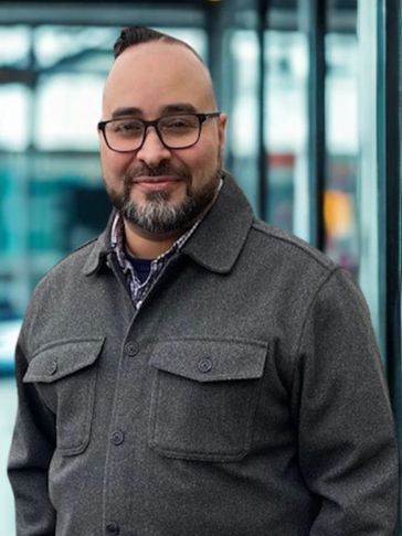 Man with glasses and a beard wearing a gray jacket, smiling outdoors.