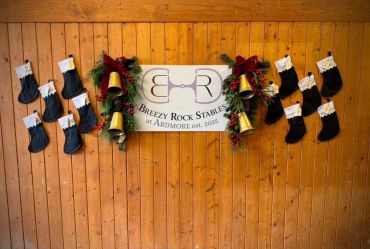 Holiday stockings and wreaths decorate a wooden wall with a Breezy Rock Stables sign.