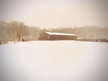 A snowy rural scene with a wooden barn and fenced area surrounded by trees.