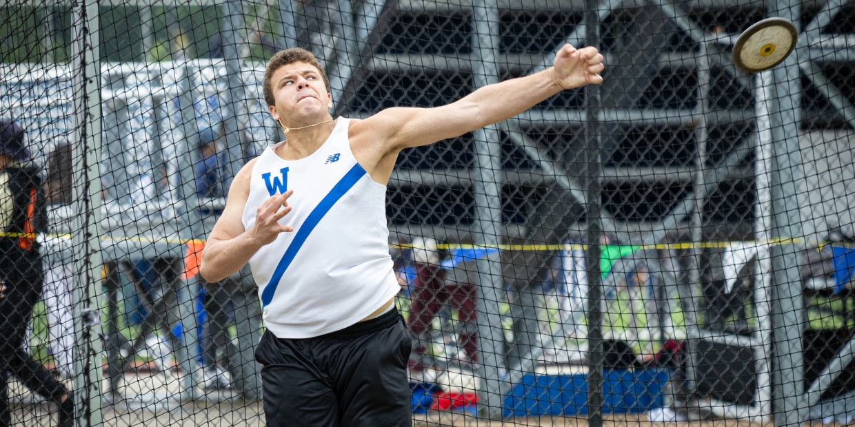 Athlete in white and blue throws a discus inside a cage during a competition.