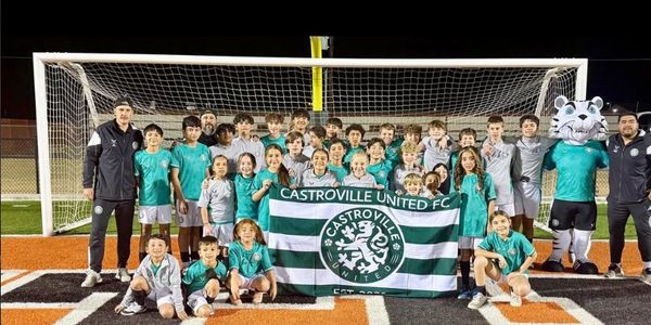 Youth soccer team posing with their club flag and mascots on a field at night.