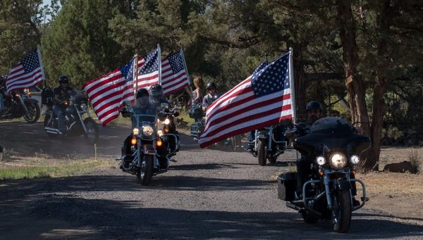 Group of motorcyclists riding with American flags on a tree-lined road.