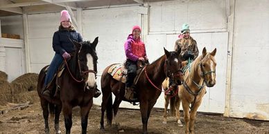 Three girls smiling on horses inside a barn with hay stacks.