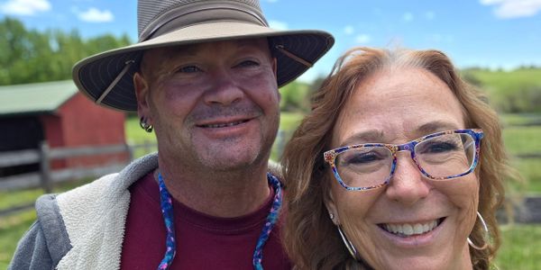 Smiling couple outdoors on a sunny day with a rustic barn in the background.