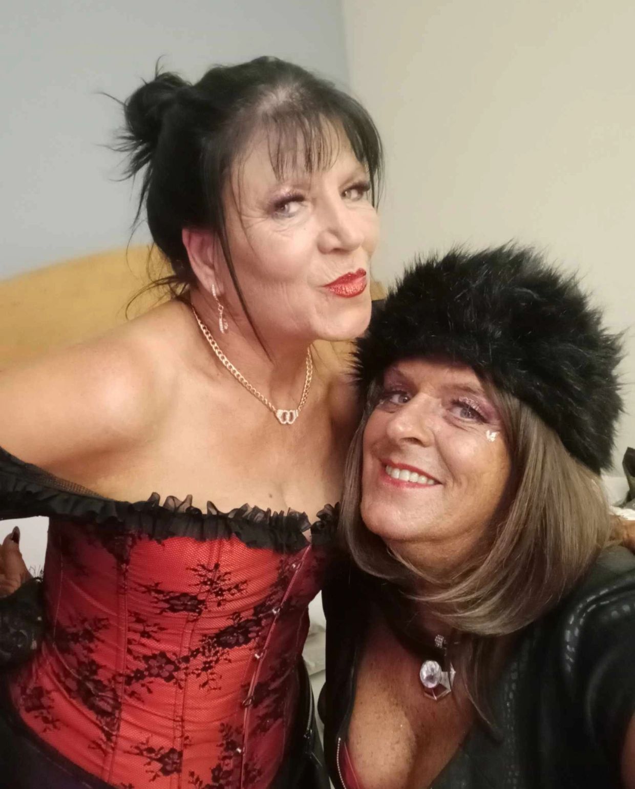 Two women posing with smiles, one in a red corset and the other in a black fur hat.