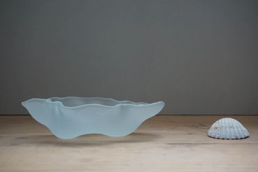 A translucent, wavy glass bowl next to a white seashell on a wooden surface.