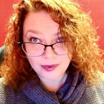 A woman with curly red hair and glasses looking at the camera.