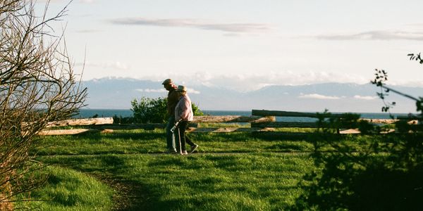 An elderly couple walks along a grassy path with mountains and ocean in the background.