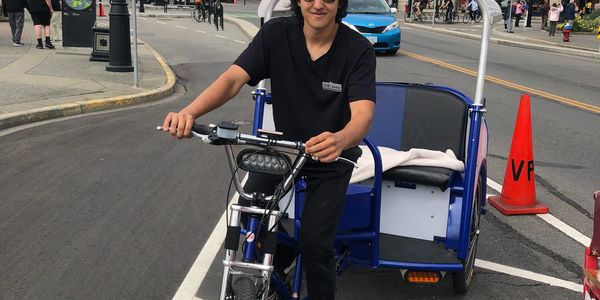 Man riding a pedicab on a city street with buildings and pedestrians.