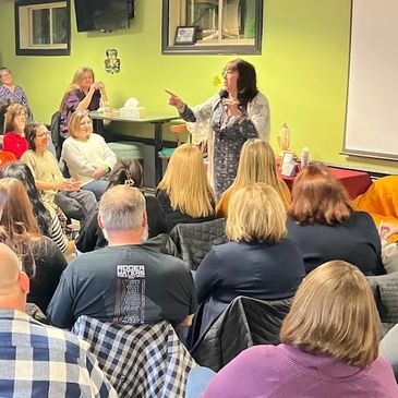 A woman speaks to an attentive audience in a cozy indoor setting.